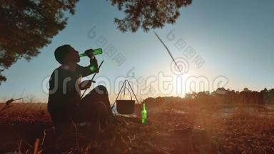 男游客坐在篝火旁喝啤酒，在大自然野营的剪影下晒日落。度假男士剪影